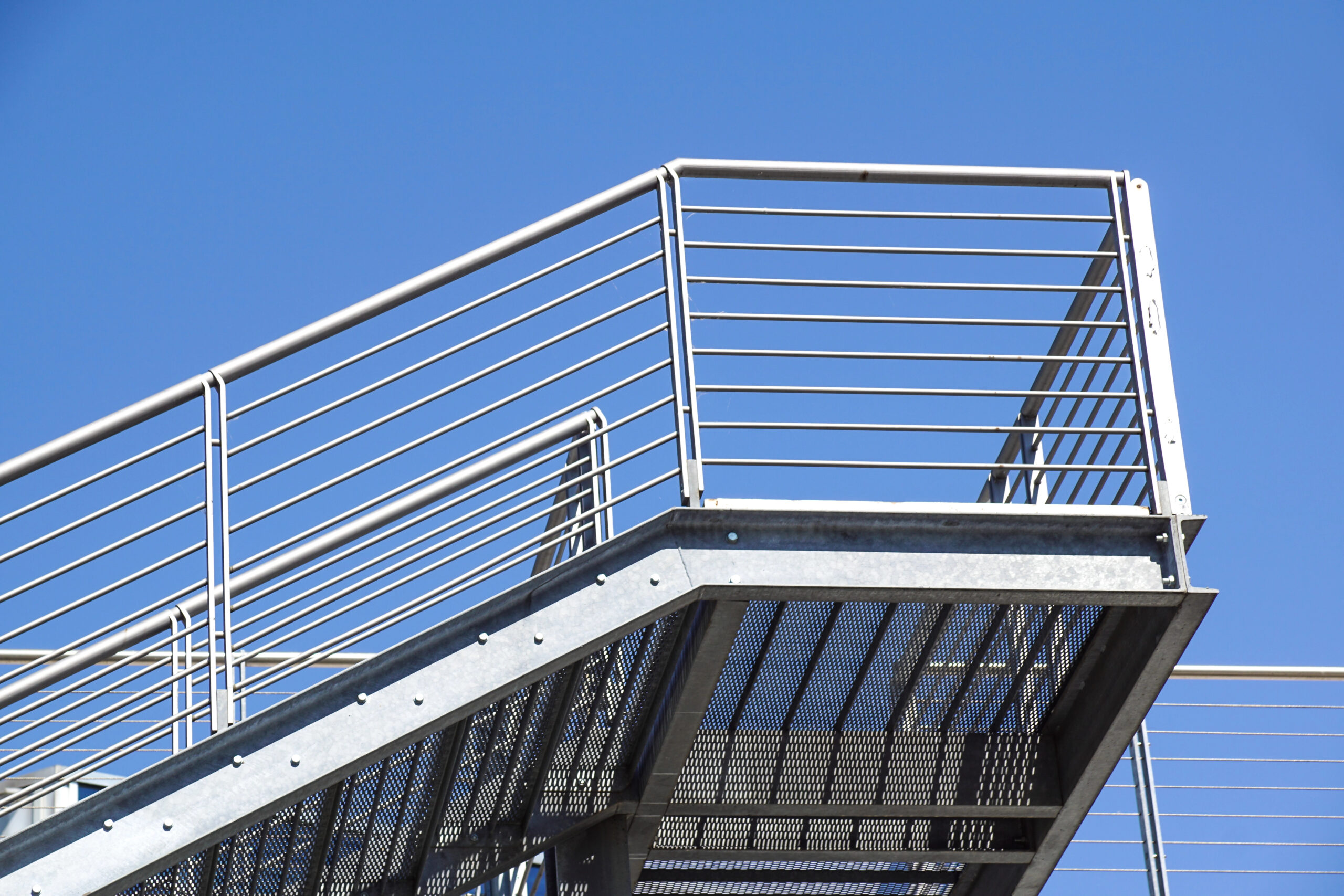 Metal staircase with custom railings and a grated platform, set against a clear blue sky. The design features horizontal bars and a sturdy steel frame, creating a modern industrial look reminiscent of sleek metal fire escapes.