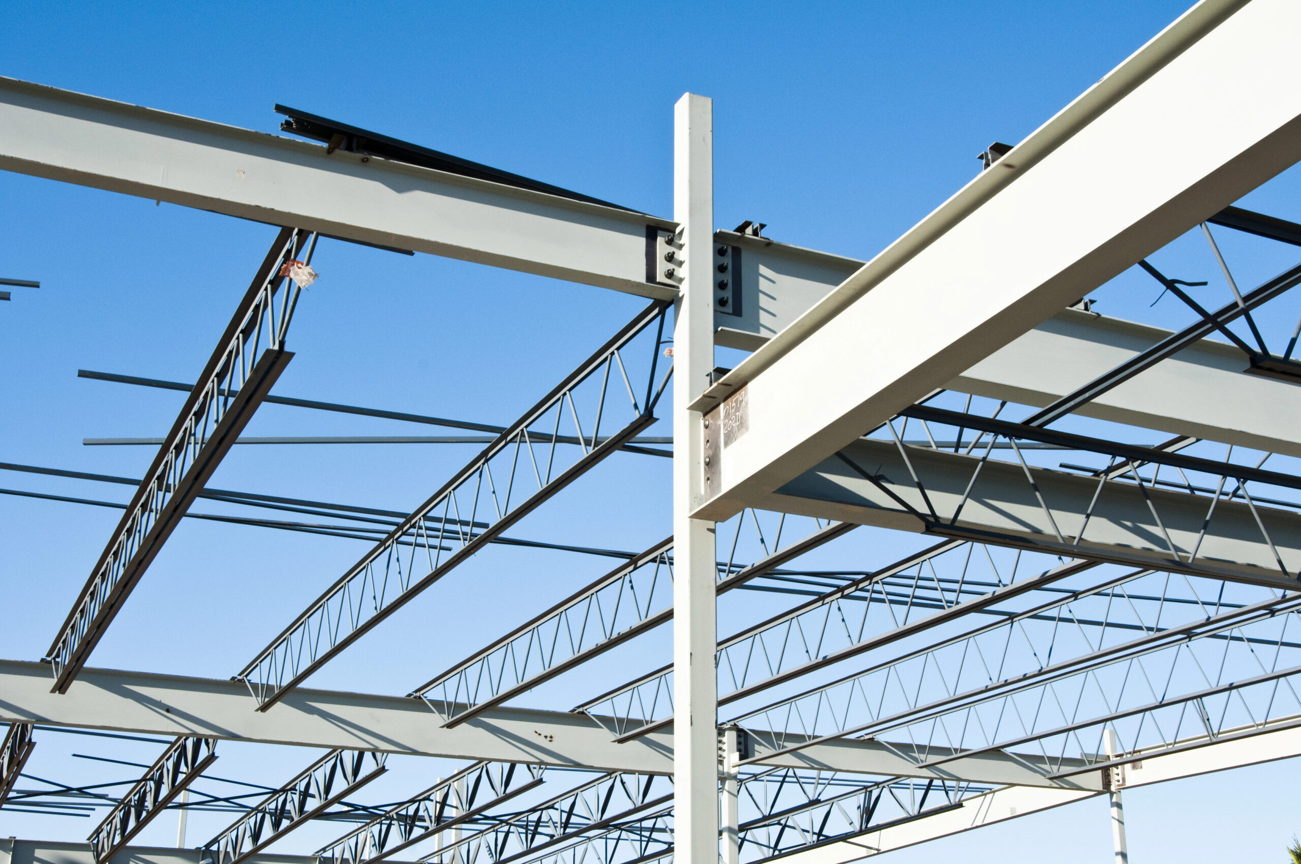 Steel beams and trusses form the skeletal structure of a building under construction, set against a clear blue sky. Nearby, custom railings are being crafted by skilled steel fabricators, indicating the early stages of architectural development.