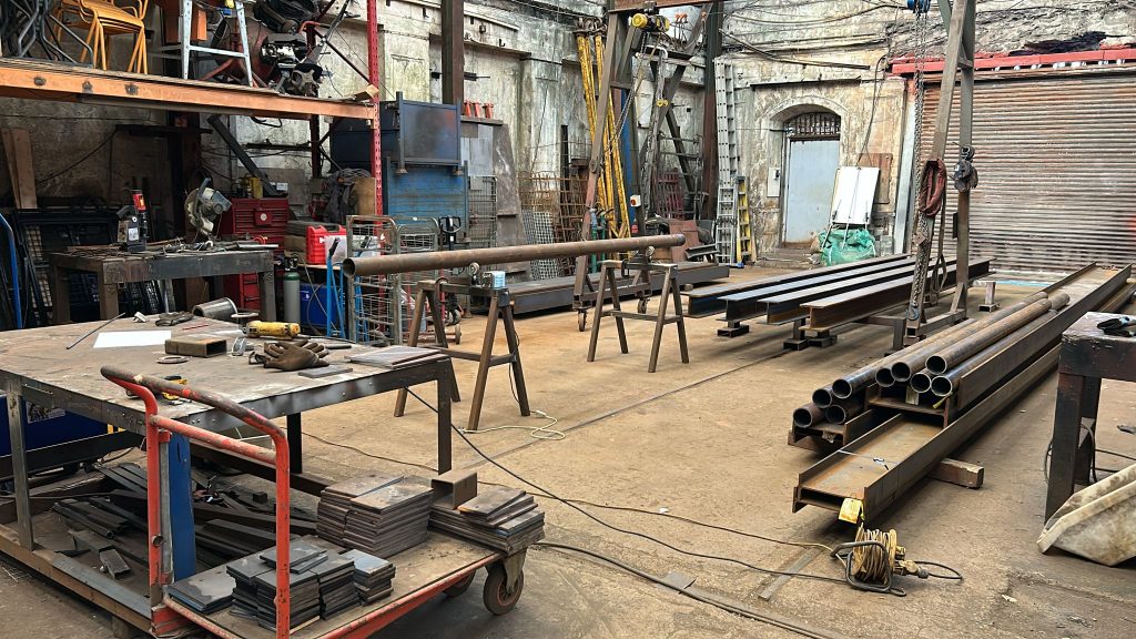 A workshop filled with metalworking tools and materials, perfect for a steel fabricator. Long metal pipes are stacked on sawhorses and tables. With a red toolbox and cutting tools scattered around, the room has a rustic, industrial vibe ideal for crafting metal fire escapes.