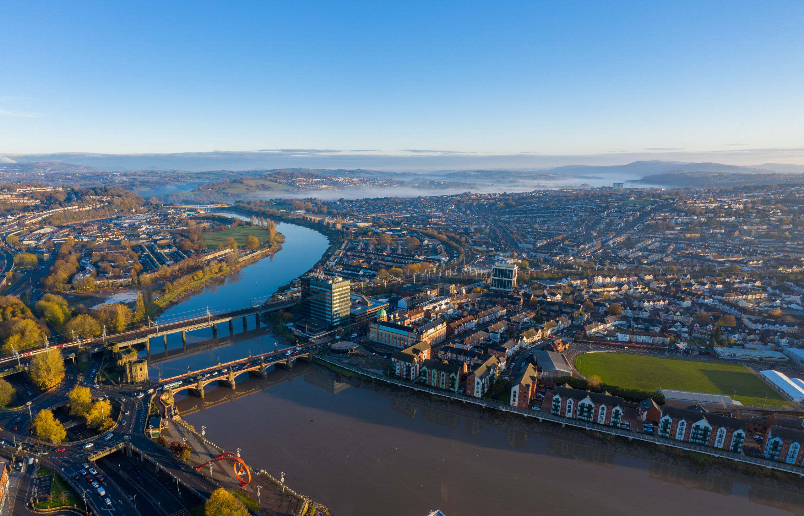 Aerial view of a city featuring a winding river, several bridges adorned with custom railings, and a mix of modern and traditional buildings. The landscape is bathed in bright sunlight, with hills visible in the background under a clear blue sky.